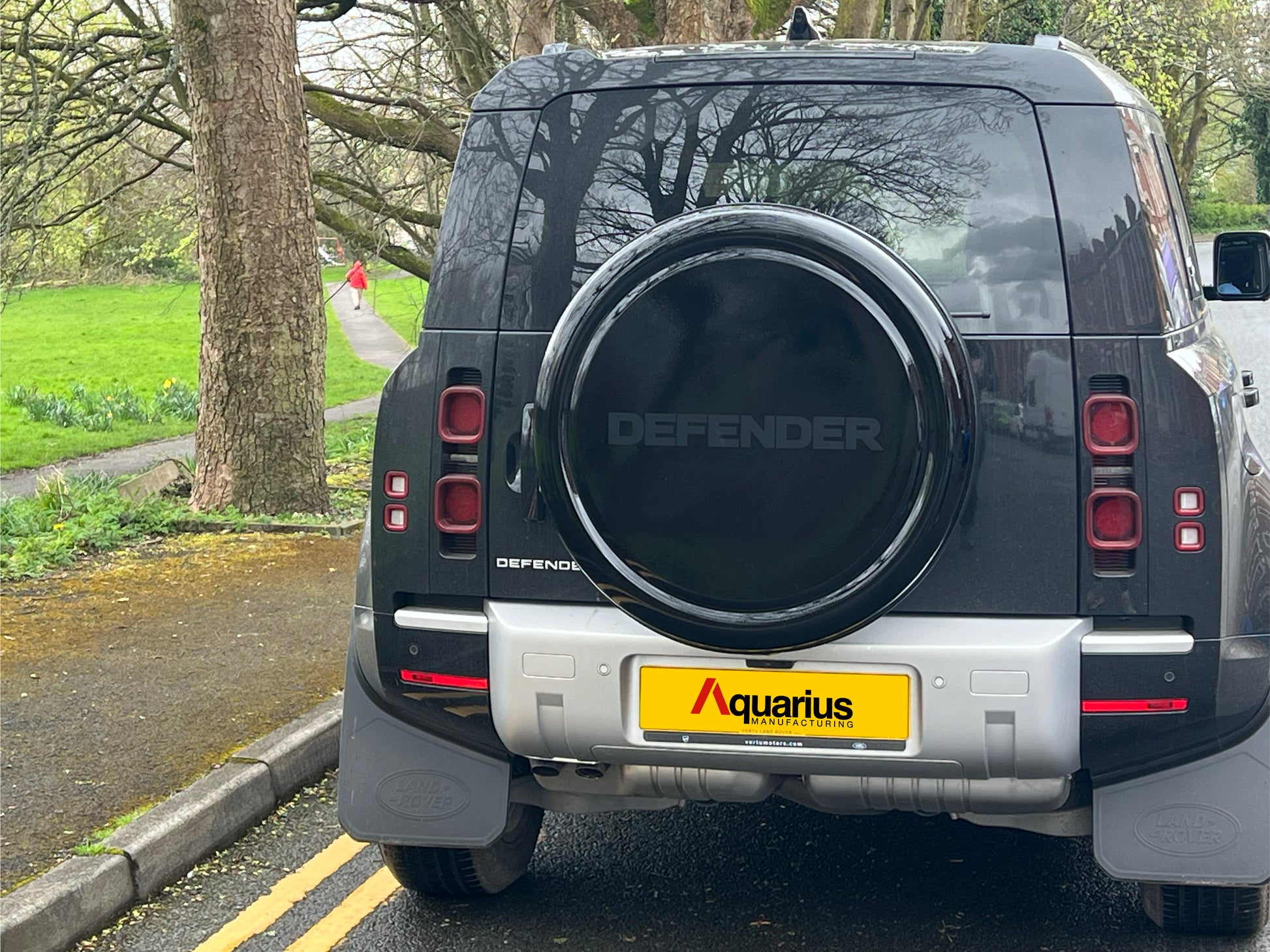 Black new shape Land Rover Defender parked next to a green park with trees, Aquarius gloss wheel cover printed "DEFENDER"