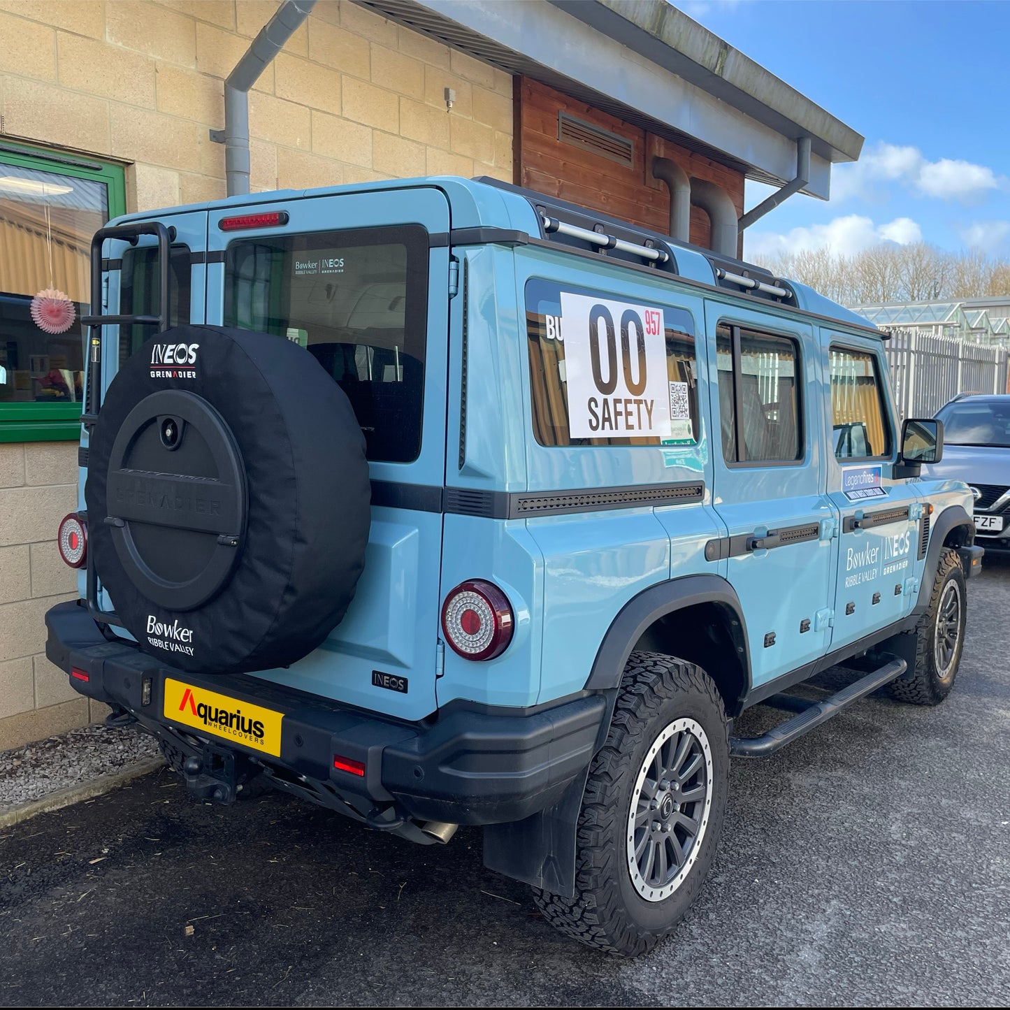 baby blue ineos grenadier with aquarius canvas wheel cover on, parked next to a building with green window frames, blue sky