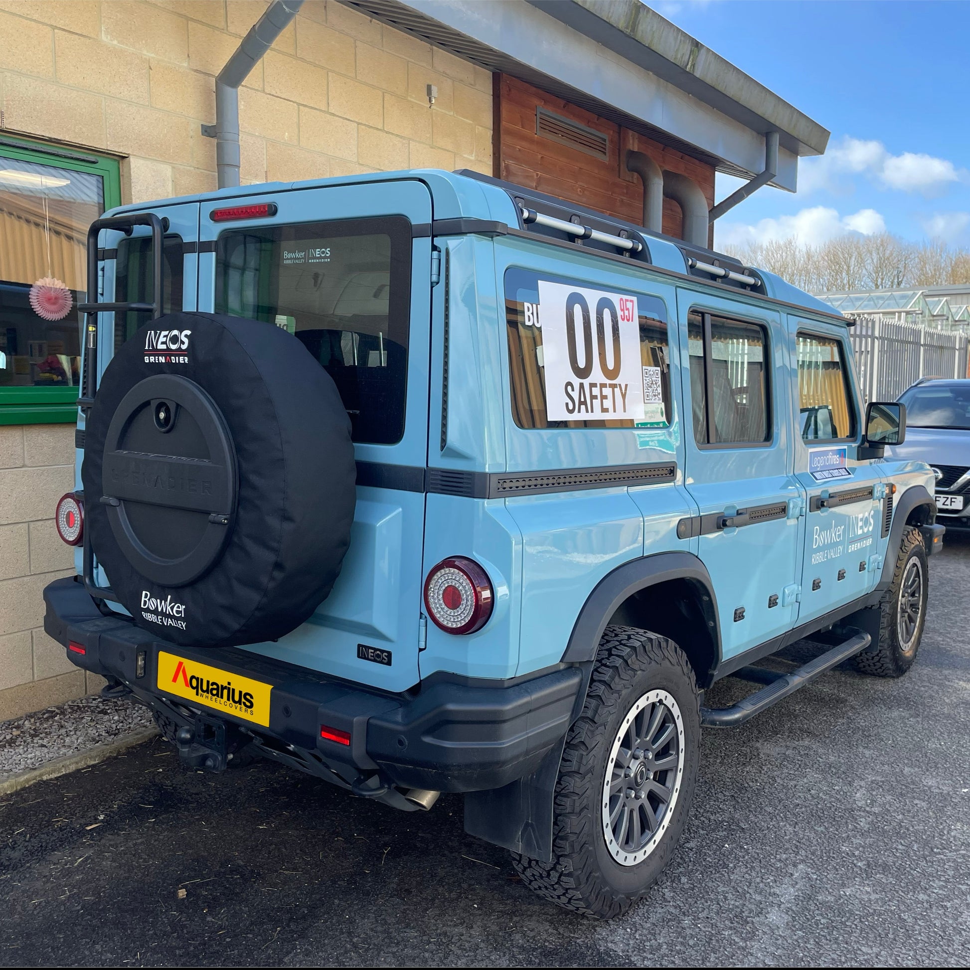 baby blue ineos grenadier with aquarius canvas wheel cover on, parked next to a building with green window frames, blue sky